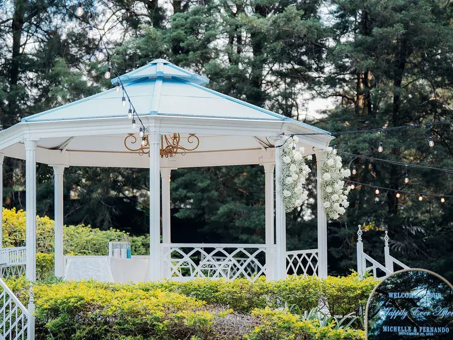 Gazebo octogonal blanco con techo azul, decorado con flores y rodeado de setos recortados, en Sala de Eventos Laureal