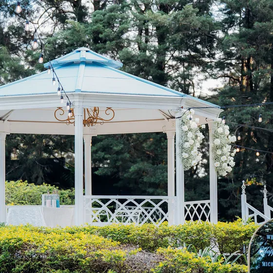 Gazebo octogonal blanco con techo azul, decorado con arco floral y rodeado de setos