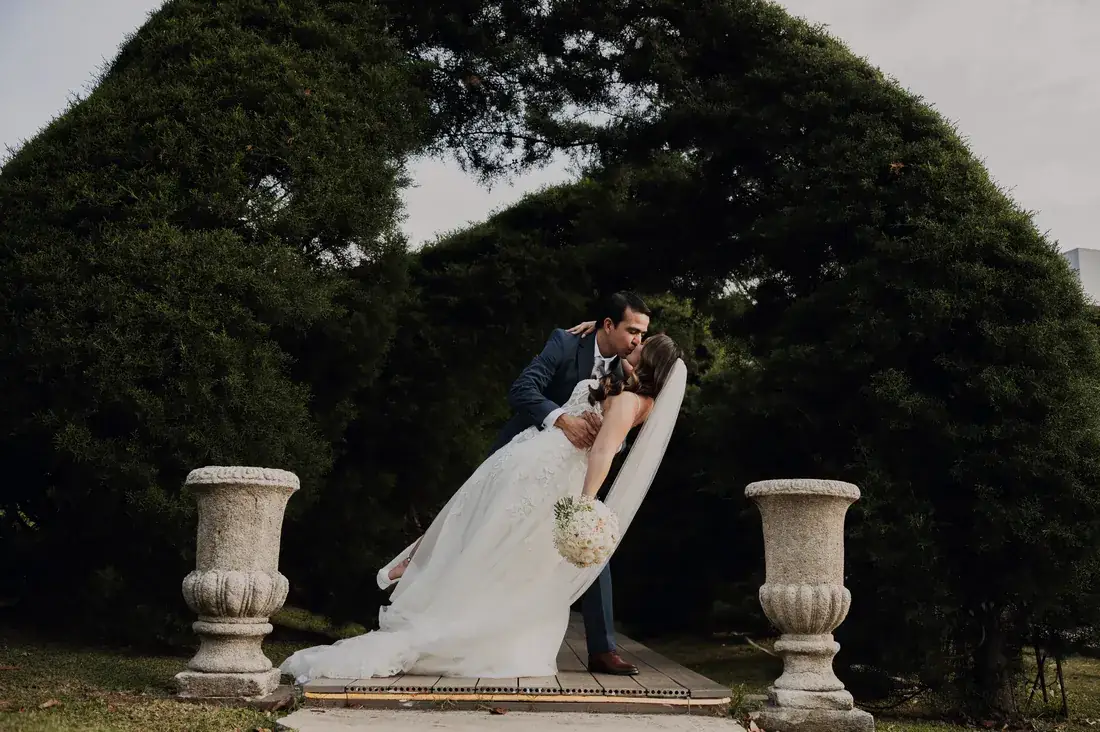 Novios besándose entre las urnas de piedra de los jardines de Sala de Eventos Laureal, bajo los árboles de la finca en San Roque de Barva, Heredia