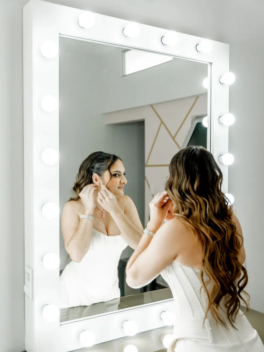 Novia colocándose los aretes frente al espejo iluminado del Salón de Maquillaje