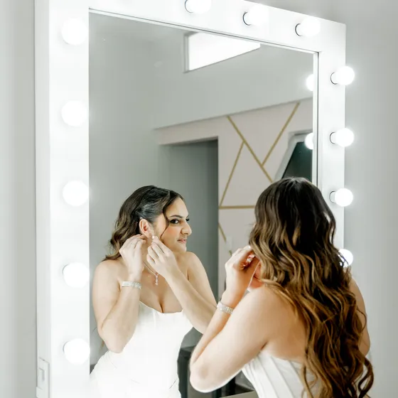 Novia colocándose los aretes frente al espejo iluminado del Salón de Maquillaje