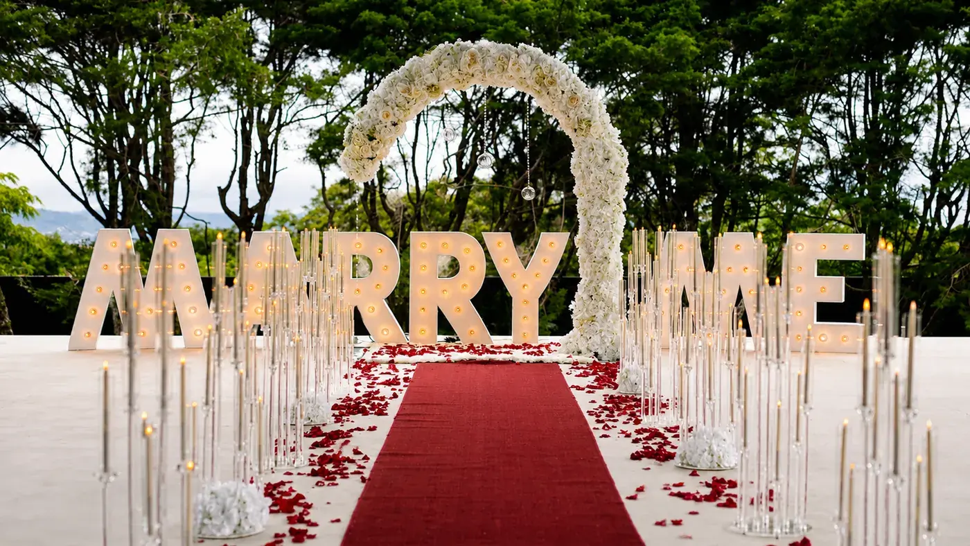 Montaje de propuesta de matrimonio en Nebulae: letras iluminadas «MARRY ME», arco floral blanco, alfombra roja con pétalos y velas