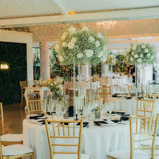 Salón montado con mesas redondas, mantelería clara y sillas tiffany doradas